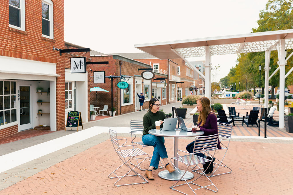 Outside a strip of shops, two women are sitting at their computers with coffees