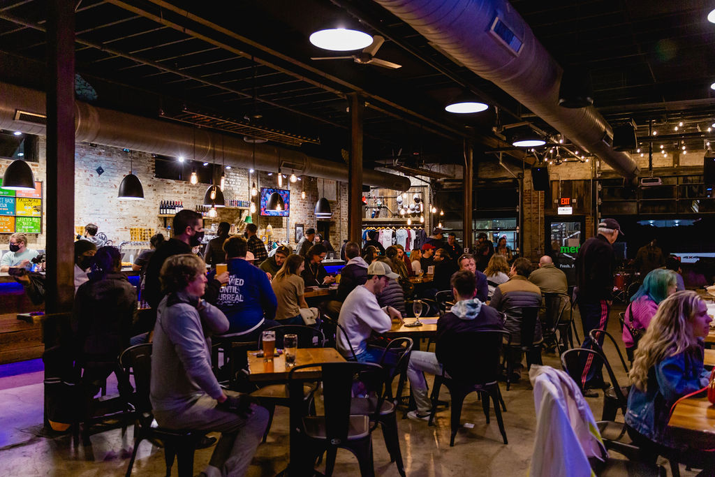 Large bar space with lots of tables that are surrounded with people drinking beer.