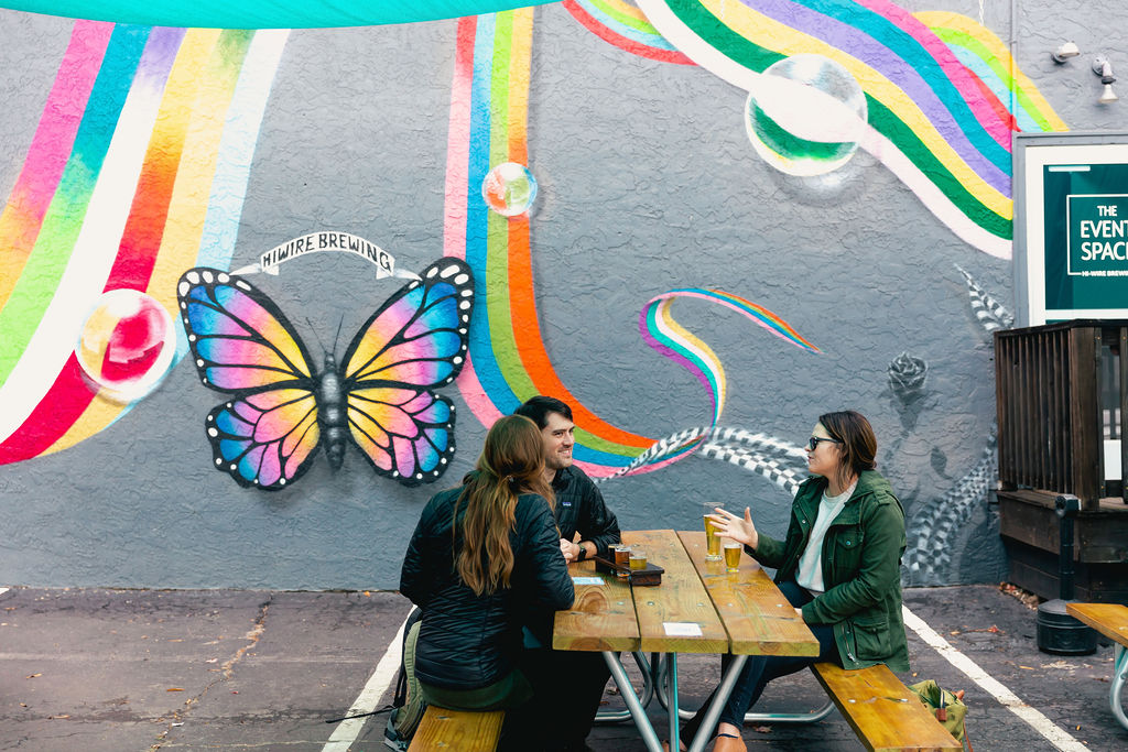 Three guests sitting at a picnic table in front of Hi-Wire's colorful butterfly mural at their Asheville location.