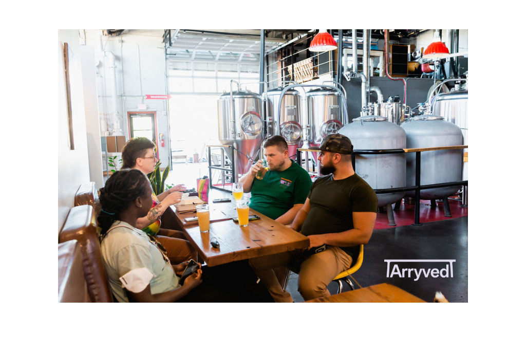 A group of 4 people sitting at a table, one drinking a beer, with beer fermenting tanks sitting behind them.