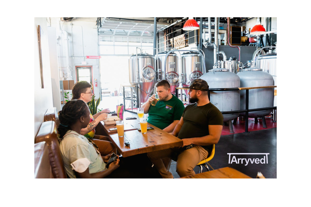 A group of 4 people sitting at a table, one drinking a beer, with beer fermenting tanks sitting behind them.